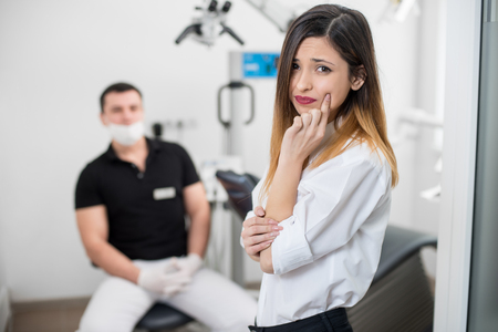 Young woman suffering from terrible teeth pain, touching cheek with hand at dental clinic. Female feeling toothache. On the background dentist sitting on the chair. Dental care and health conceptの写真素材