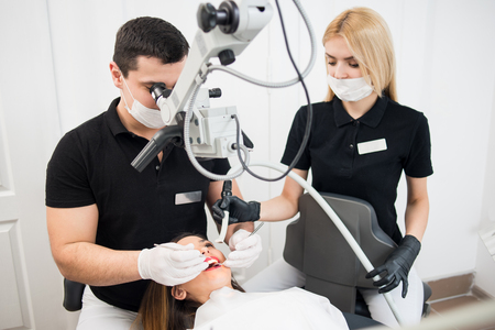 Male dentist and female assistant treating patient teeth with dental tools - microscope, mirror and probe at dental clinic office. Medicine, dentistry and health care concept. Dental equipmentの写真素材
