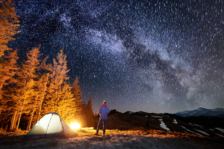 Male tourist have a rest in his camp near the forest at night. Man standing near campfire and tent under beautiful night sky full of stars and milky way, and enjoying night scene. Long exposureの写真素材