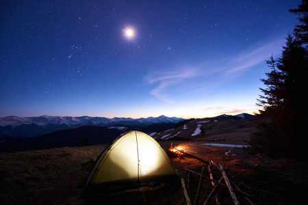Tourist camping near forest in the mountains. Illuminated tent and campfire under beautiful evening sky full of stars and the moonの写真素材