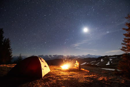 Male hiker have a rest in his camp near the forest at night. Man sitting near campfire and tent under beautiful night sky full of stars and the moon, and enjoying night scene in the mountainsの写真素材