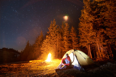 Male tourist have a rest in his camp near the forest at night. Man sitting near campfire and tent under beautiful night sky full of stars and enjoying night sceneの写真素材