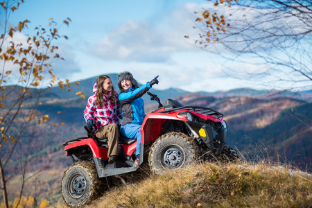 Woman is showing something in distance to her friend in nature on the autumn sunny day. Two girlfriends in winter clothing on a red ATV at a hill on the background of mighty mountains and blue skyの写真素材
