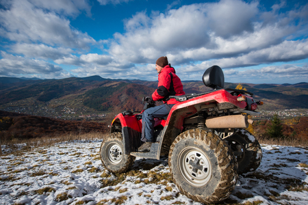 Sports ATV quad bike with rider at the snow-covered slope against the blue cloudy sky, mountains and the town in the valley on a sunny day in the mountainsの写真素材
