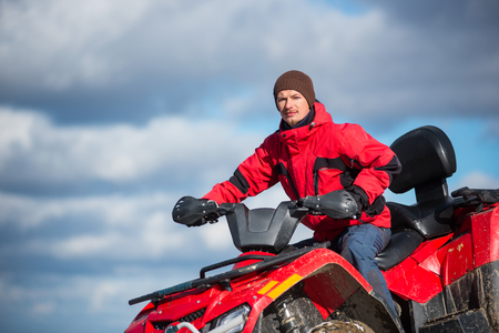 Close-up portrait of man in a red winter clothes riding on red quad bike in the mountains top. Blue sky with clouds on the backgroundの写真素材