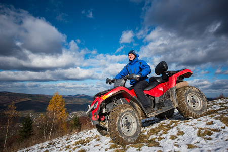 Smiling male on the quad riding down on the snowy slope on a sunny autumn day. Beautiful mountain landscape under a blue cloudy sky with the town in the valleyの写真素材