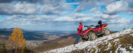 Panorama. Man standing near ATV quad bike, looking into the distance on snowy mountain top. On the background blue cloudy sky, mountains and the town in the valley with copy space. Autumnの写真素材