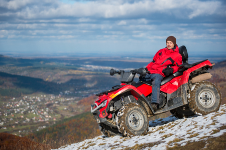 Man in winter clothes resting on a red atv bike on a snowy mountain top, looking to the camera on a blurred background of mountains and the town in the valley with copy spaceの写真素材