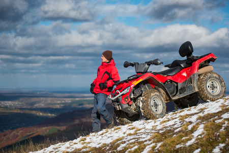Atv quad bike near man in winter clothes looks into the distance on snowy mountain slope. On the background blue cloudy sky, mountains and the town in the valley with copy spaceの写真素材