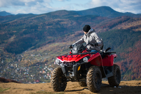 Man in winter clothes on a red quad bike on a mountain top enjoying the view of the mighty mountains behind him. Blurred background of mountain sceneryの写真素材
