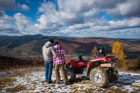 Rear view of a guy hugging a girl near the ATV on a mountain top under the blue sky with cumulus clouds at background of mighty mountains and village in the valleyの写真素材