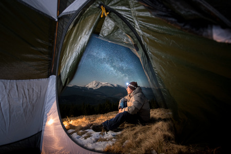 View from inside a tent on the male tourist have a rest in his camping in the mountains at night. Man with a headlamp sitting near campfire under beautiful night sky full of stars and milky wayの写真素材