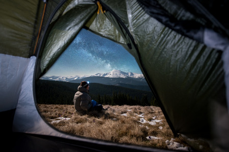 View from inside a tent on the male hiker have a rest in his camping at night. Man with a headlamp sitting under beautiful night sky full of stars and milky way. Snow-covered mountains on backgroundの写真素材