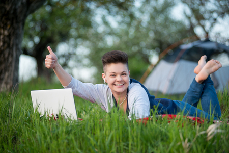 Young woman smiling to the camera, lying on the grass with her laptop, showing thumbs up to the camera student working internet online education travelling wireless connectionの写真素材