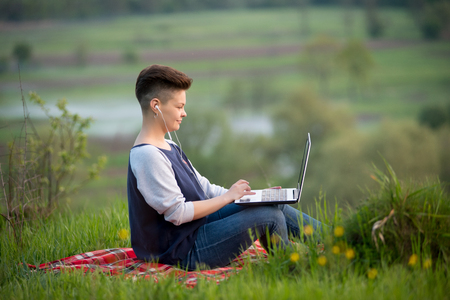Young woman wearing headset while working on her laptop sitting on the grass outdoors copyspace technology online connectivity mobility wireless internet projectの写真素材