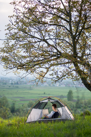 Vertical shot of a woman sitting in a tent on top of a hill under tree using her laptop while camping lifestyle active online connectivity mobility technologyの写真素材