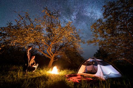 Young couple tourists standing at a campfire near tent under trees and beautiful night sky full of stars and milky way. Night camping. Long exproseの写真素材