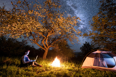 Female tourist using her laptop with headset in the camping at night. Woman sitting near campfire and tent under trees and beautiful night sky full of stars and milky wayの写真素材
