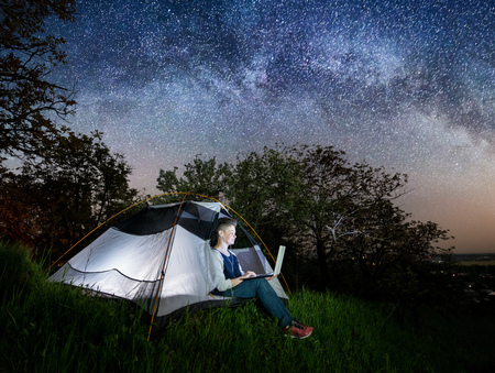 Woman using her laptop with headset in the camping at night. Woman sitting in the tent under beautiful night sky full of stars and milky wayの写真素材