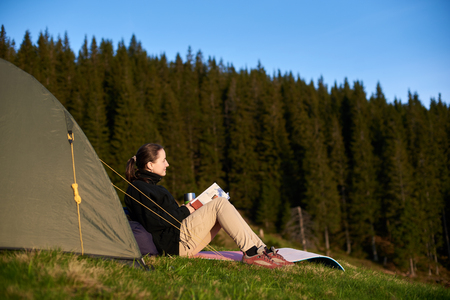 Young happy female hiker enjoying in the camping with book and cup of coffee near the tent in the eveningの写真素材
