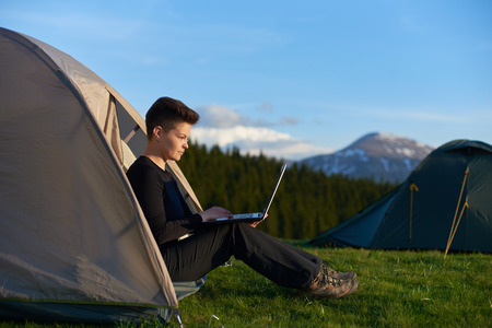 Young female tourist enjoying sunrise in the mountains sitting in her tent working on the laptop copyspace camping site travelling mobility technology internet online studying tourism.の写真素材