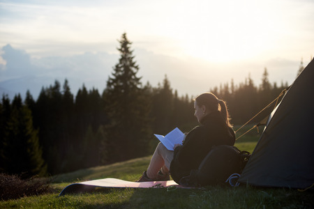 Young woman hiker reading a book near the tent in the evening at sunsetの写真素材