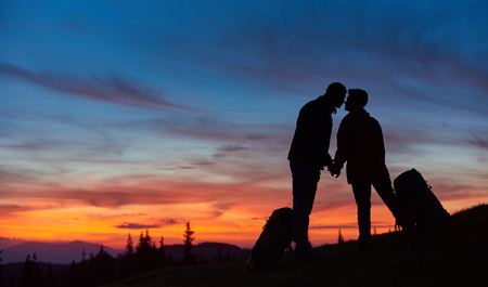 Silhouettes of a loving couple of hikers kissing on top of the mountain holding hands standing near their backpacks copyspace love anniversary achieving togetherness affection romantic hiking active.の写真素材