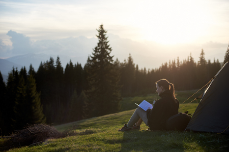 Young woman tourist sitting near the tent and reading a book at sunset in the mountainsの写真素材
