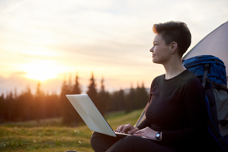 Happy woman looking away thoughtfully using her laptop outdoors while camping sitting near her tent dreaming achievement success travel online connectionの写真素材