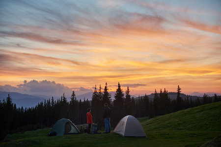 Man and woman having a rest in the camping near two tents in the mountains while hiking together with their backpacks enjoying beautiful sunset with fiery colorful sky love relationships nature.の写真素材
