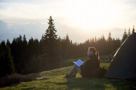 Young woman backpacker having a rest with cup of coffee and book near the tent in the evening at sunsetの写真素材
