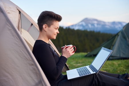 Shot of a young female relaxing in her tent on the camping site holding a cup looking away thoughtfully after working on her laptopの写真素材