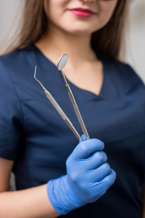 Dentist with blue gloves holding tools - dental mirror and dental probe at the dental office. Close-up, selective focus on tools. Dentistryの写真素材
