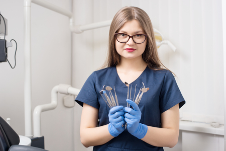 Portrait of young female attractive dentist holding dental tools - mirrors and probes at the morden dental office. Doctor wearing glasses, blue uniform, gloves and looking to the camera. Dentistryの写真素材