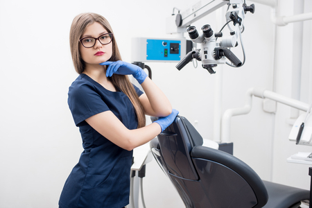 Portrait of beautiful female dentist at the modern dental office. Doctor wearing glasses, blue uniform, blue gloves and looking to the camera. Dentistryの写真素材