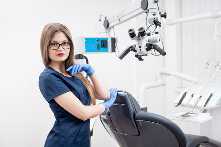 Portrait of young female dentist at the morden dental clinic. Doctor wearing glasses, blue uniform, blue gloves. Dentistryの写真素材