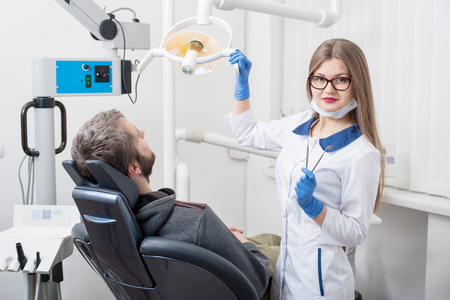 Friendly female dentist holding dental tools - lamp, mirror and probe and getting ready for dental procedure with male patient in modern dental clinic. Doctor looking to the camera. Dentistryの写真素材