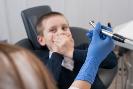 Dentist in gloves holding in hand dental drill bit and kid frightened by dentists covers her mouth. The hand dental with drill bit is in focusの写真素材