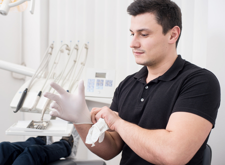 Handsome male dentist is wearing white gloves at the dental office. Stomatology equipment on the backgroundの写真素材