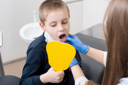 A boy in dentist's chair looks in the mirror and the doctor in blue gloves shows him his teeth.の写真素材