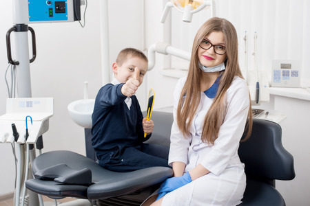 A boy smiling in dentist's chair and showing thumbs up gestures of good class. Doctor and child looks at camera.の写真素材