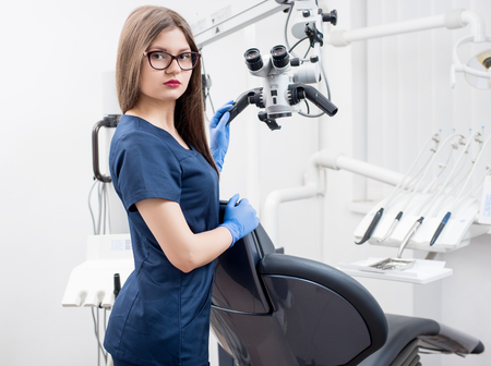 Portrait of beautiful female dentist with microscope at the morden dental office. Doctor wearing glasses, blue uniform, blue gloves and looking to the camera. Dentistryの写真素材