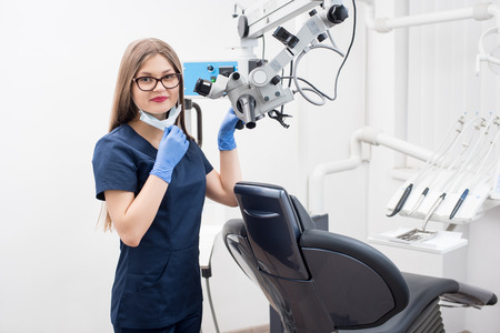 Portrait of beautiful female dentist with microscope at the morden dental office. Doctor wearing mask, glasses, blue uniform, blue gloves and looking to the camera. Dentistryの写真素材