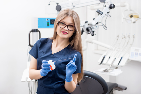 Portrait of young smiling female dentist with dental floss in the dental office. Doctor wearing blue uniform and gloves, glasses. Dentistry. Stomatology equipment on the backgroundの写真素材