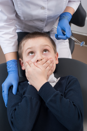 Boy frightened by dentists covers her mouth and looks for him. Doctor in gloves with one hand holds the instruments with the other hand supports the patientの写真素材