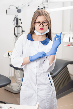 Portrait of young beautiful female dentist wearing blue gloves, glasses, white uniform and looking to the camera at the morden dental office. Dentistryの写真素材