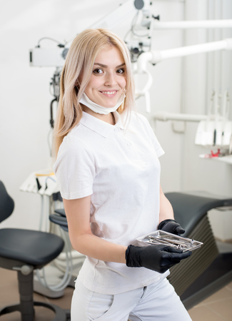 Portrait of young attractive female dentist holding dental instruments at the morden dental office. Doctor wearing mask, black gloves and looking to the camera. Dentistryの写真素材