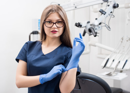 Close-up portrait of beautiful female dentist at the morden dental office. Doctor wearing glasses, blue uniform, blue gloves. Dentistryの写真素材