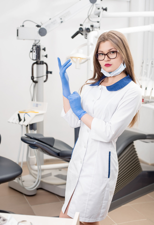 Portrait of young attractive female dentist wearing blue gloves, glasses, mask, white uniform at the morden dental office. Dentistryの写真素材