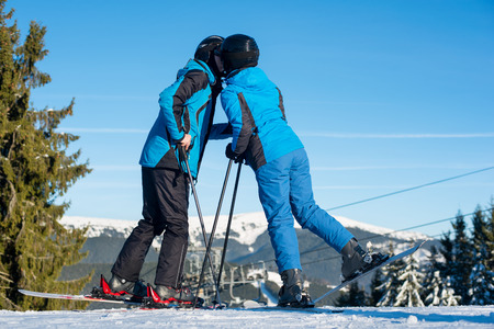 Couple skiers kissing on mountain top with mountain and blue sky in background. Dressed at blue warm clothing and helmets. Sunny winter dayの写真素材